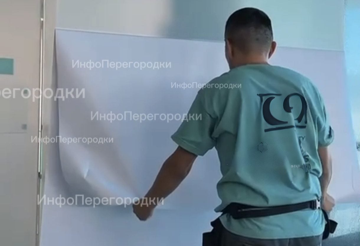 Мастер со спины, плёнка закрывает стекло почти на всю ширину Оклейка стекла плёнкой: мастер раскатывает светлое полотно по остеклению