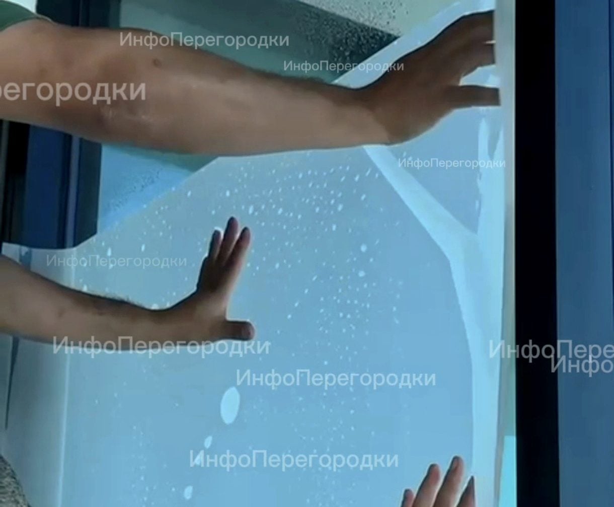 Две руки разглаживают плёнку на остеклении в тёмном профиле Плёнку прикатывают к стеклу ладонями: под ней ещё видны капли воды